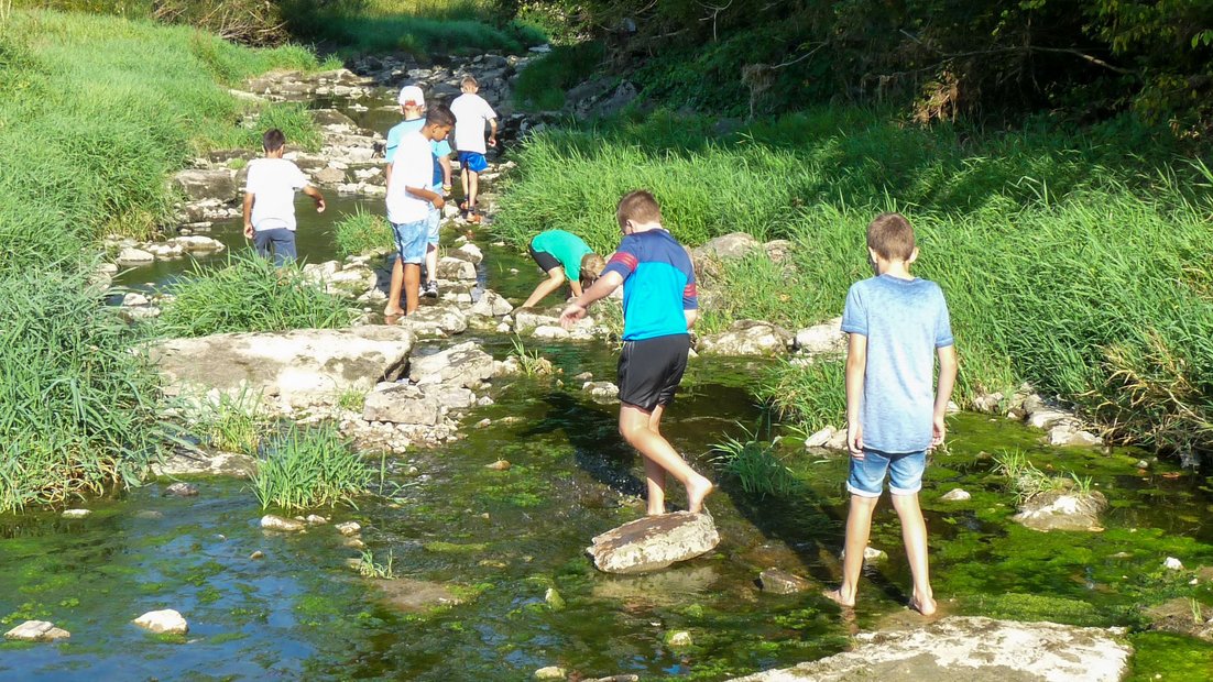 Младшие школьники исследуют мелкое русло реки с большими камнями во время дня походов.